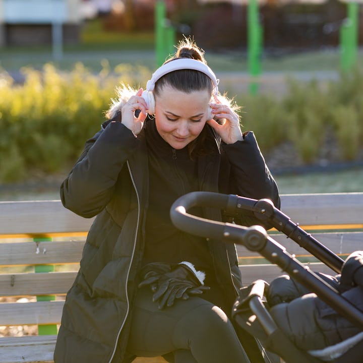 A woman sitting on a bench in a public park with her baby in a pram. She is wearing white headphones...
