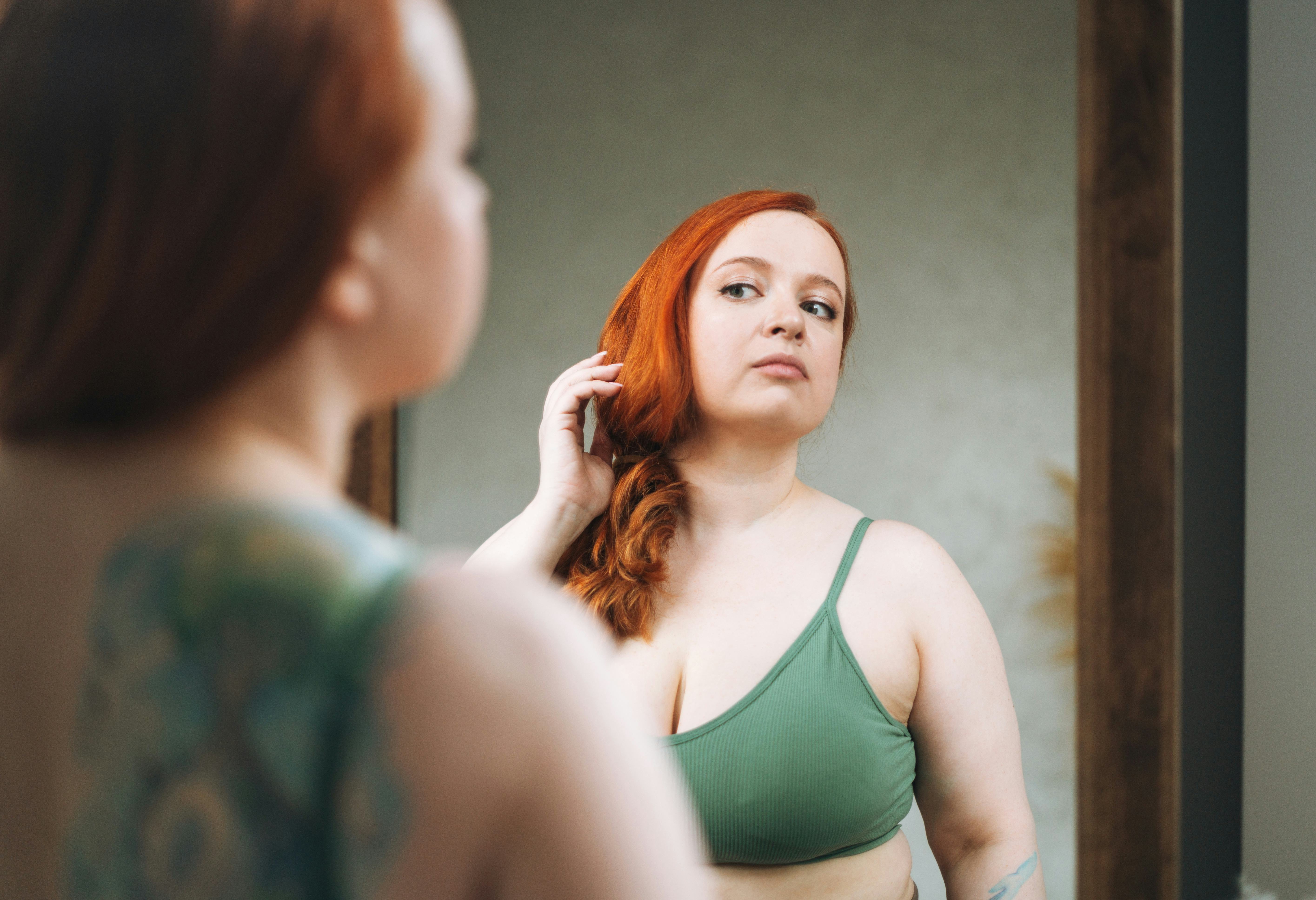 Self loving red haired plus size young woman in underwear looking at mirror in bedroom at home