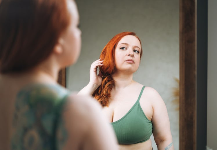 Self loving red haired plus size young woman in underwear looking at mirror in bedroom at home
