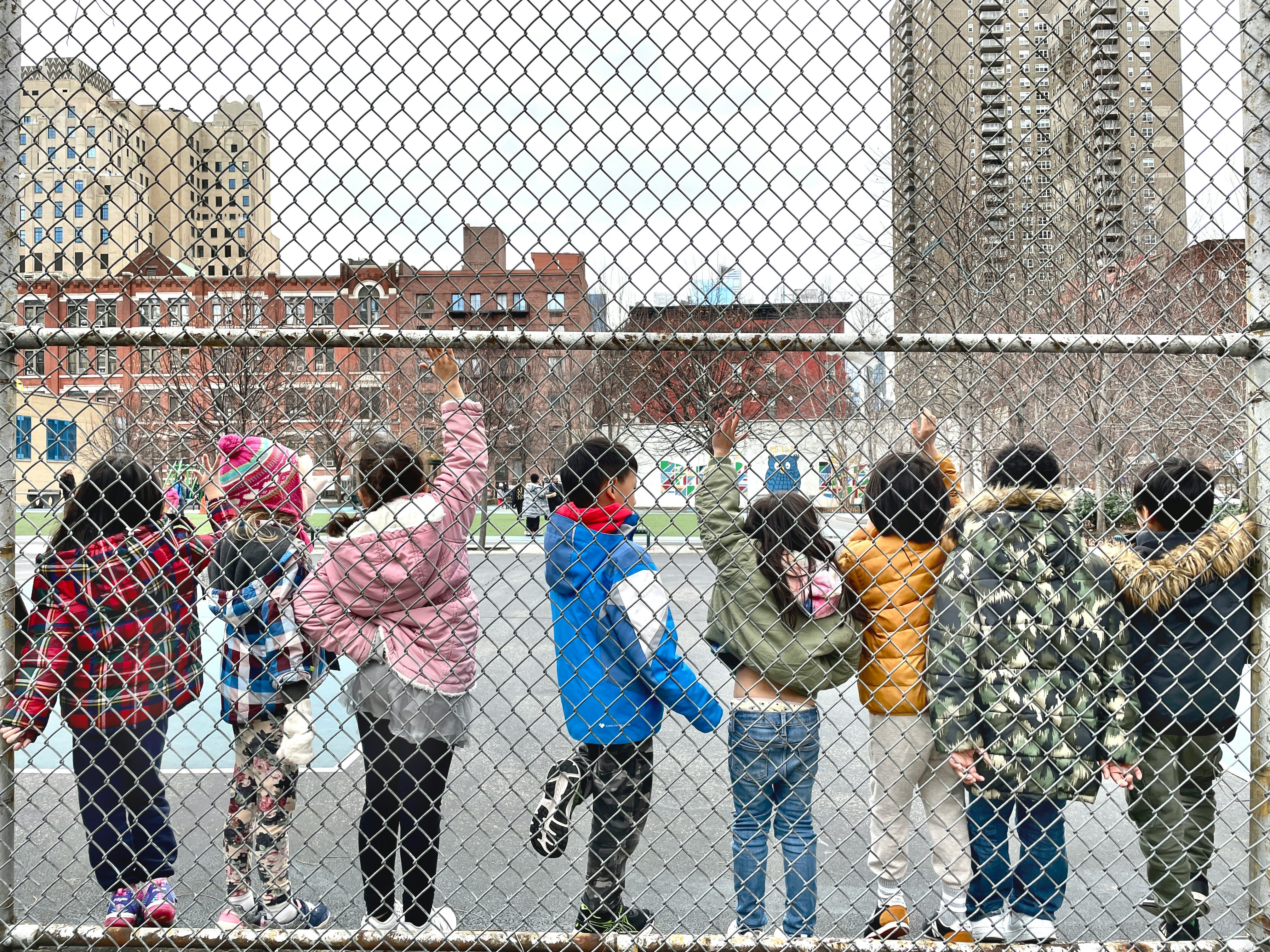 A row of students ages 5-7 raising their hands while standing along a chain-link fence on a playgrou...