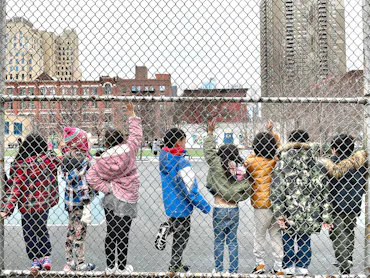 A row of students ages 5-7 raising their hands while standing along a chain-link fence on a playgrou...