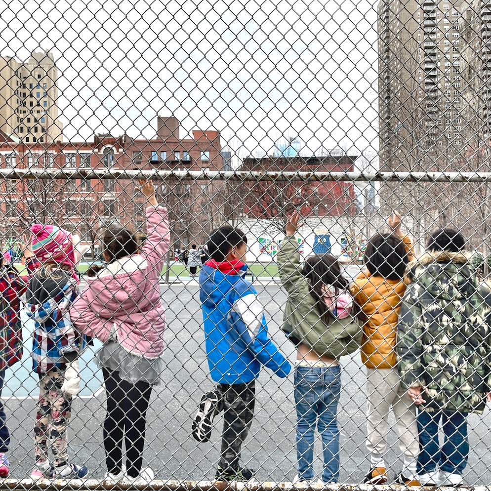 A row of students ages 5-7 raising their hands while standing along a chain-link fence on a playgrou...