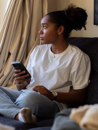 A teen sitting on a couch and looking out a window while playing on her phone.