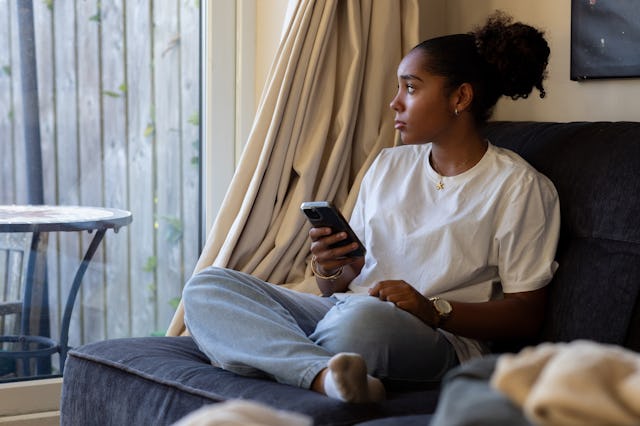 A teen sitting on a couch and looking out a window while playing on her phone.