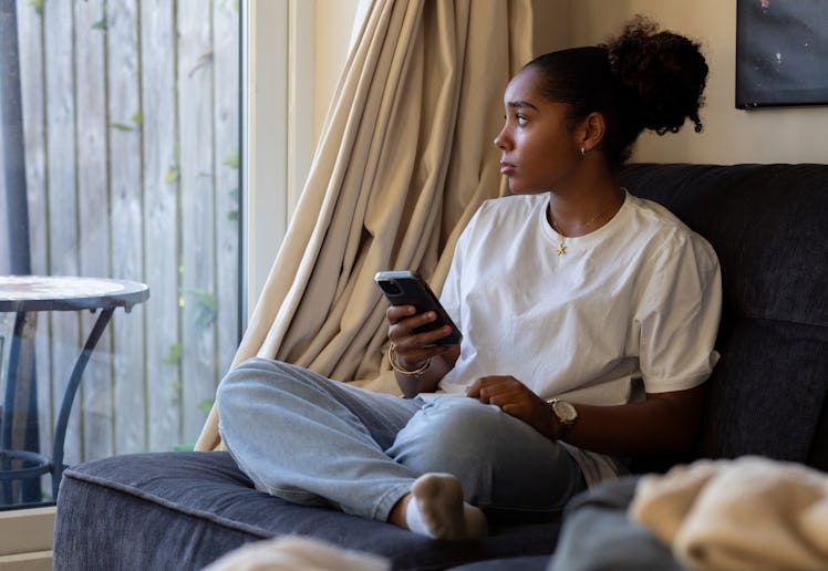 A teen sitting on a couch and looking out a window while playing on her phone.