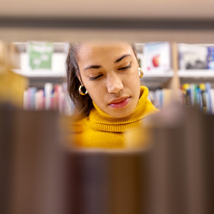 African woman student looking for book at library bookshelf. Young female searching for book on book...