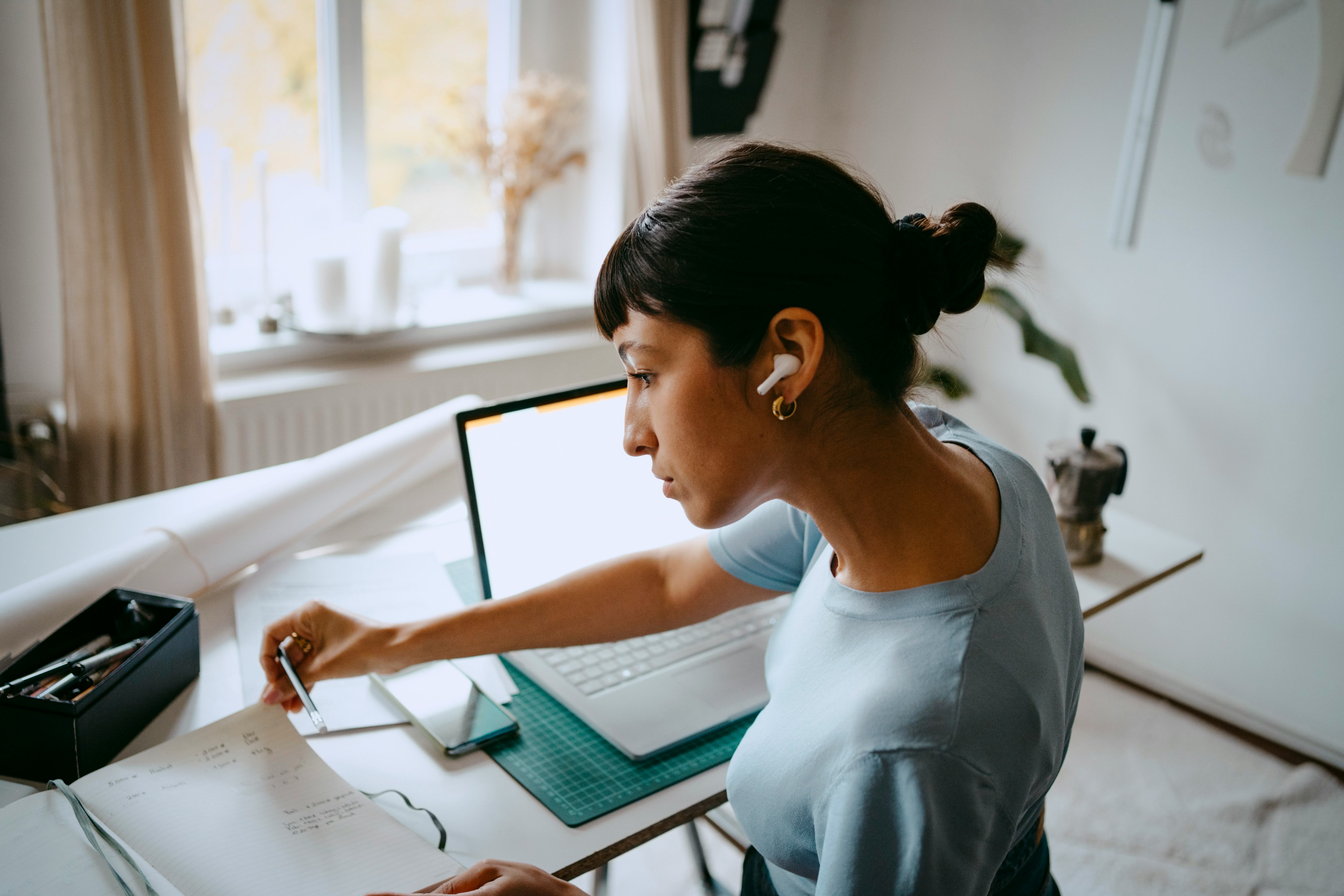 A woman sits at a desk with ear buds in, flipping through a notebook.