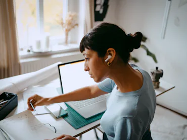 A woman sits at a desk with ear buds in, flipping through a notebook.