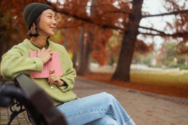 Smiling woman with cap and green sweater holding a book in her hands. She is delighted with the book...