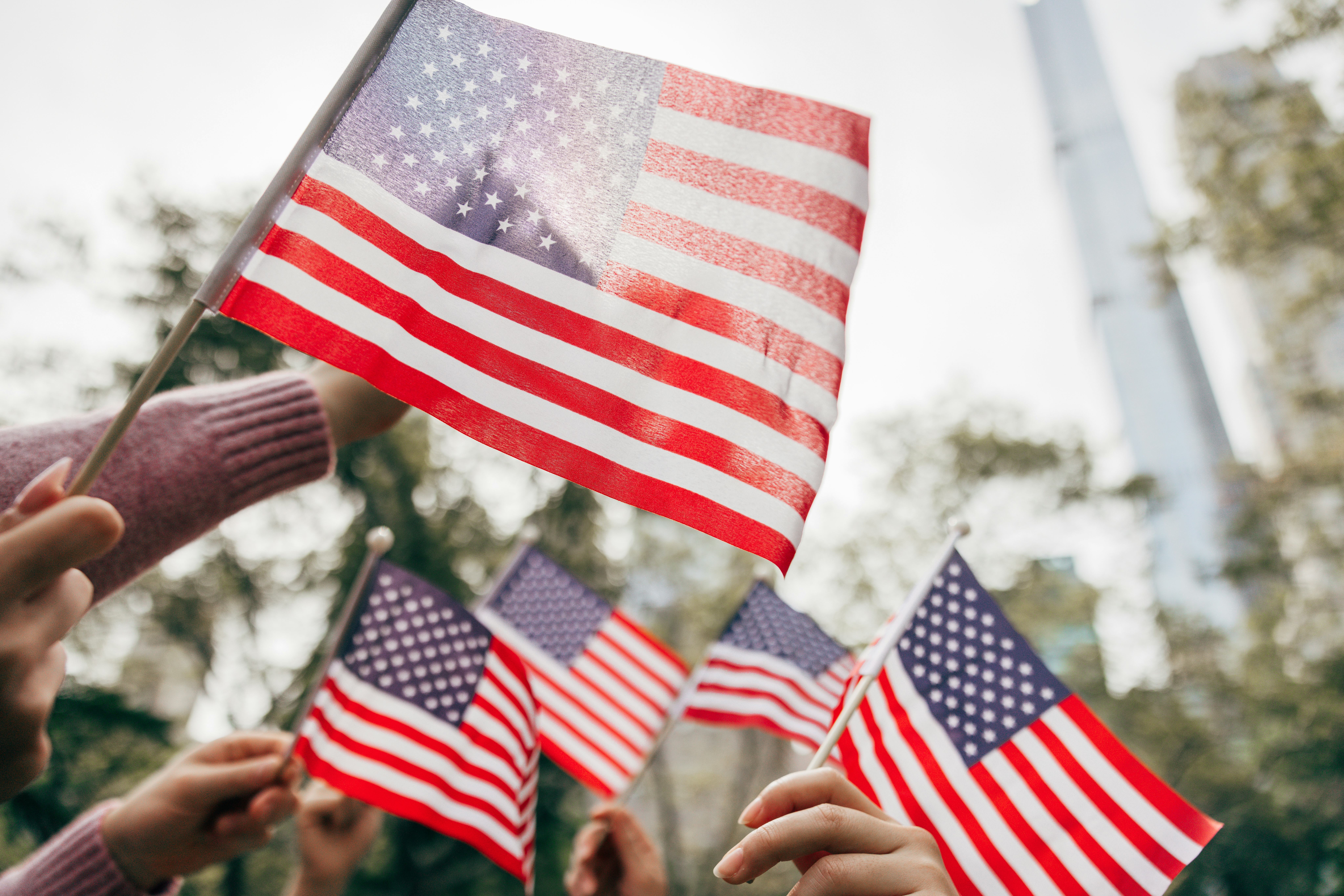 Hands grasping American flags reach upward in a collective display of patriotism, set against a back...