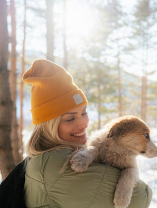 A woman in a warm green coat holds a dog as they enjoy a winter day in the woods.