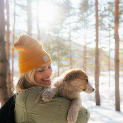 A woman in a warm green coat holds a dog as they enjoy a winter day in the woods.