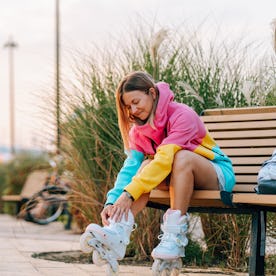 Young woman sitting on a bench and putting on her roller skates, getting ready for rollerblading in ...
