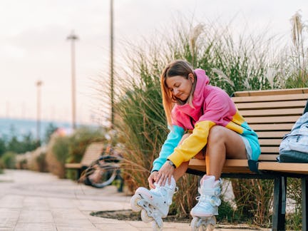 Young woman sitting on a bench and putting on her roller skates, getting ready for rollerblading in ...