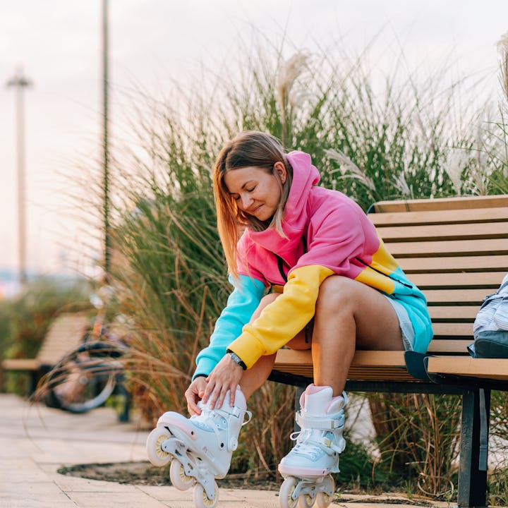 Young woman sitting on a bench and putting on her roller skates, getting ready for rollerblading in ...