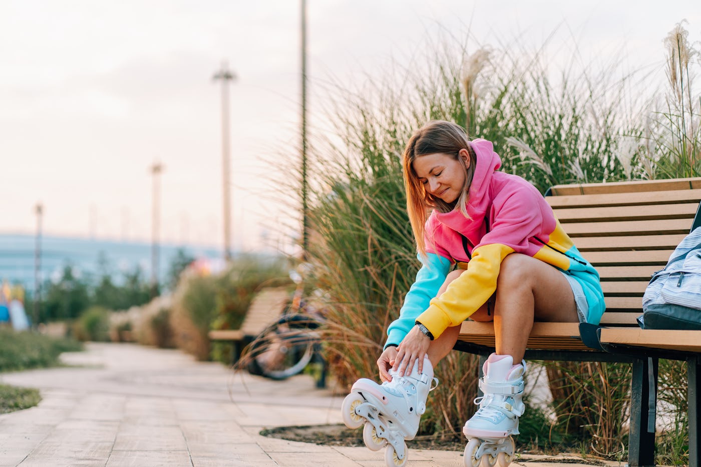 Young woman sitting on a bench and putting on her roller skates, getting ready for rollerblading in a park