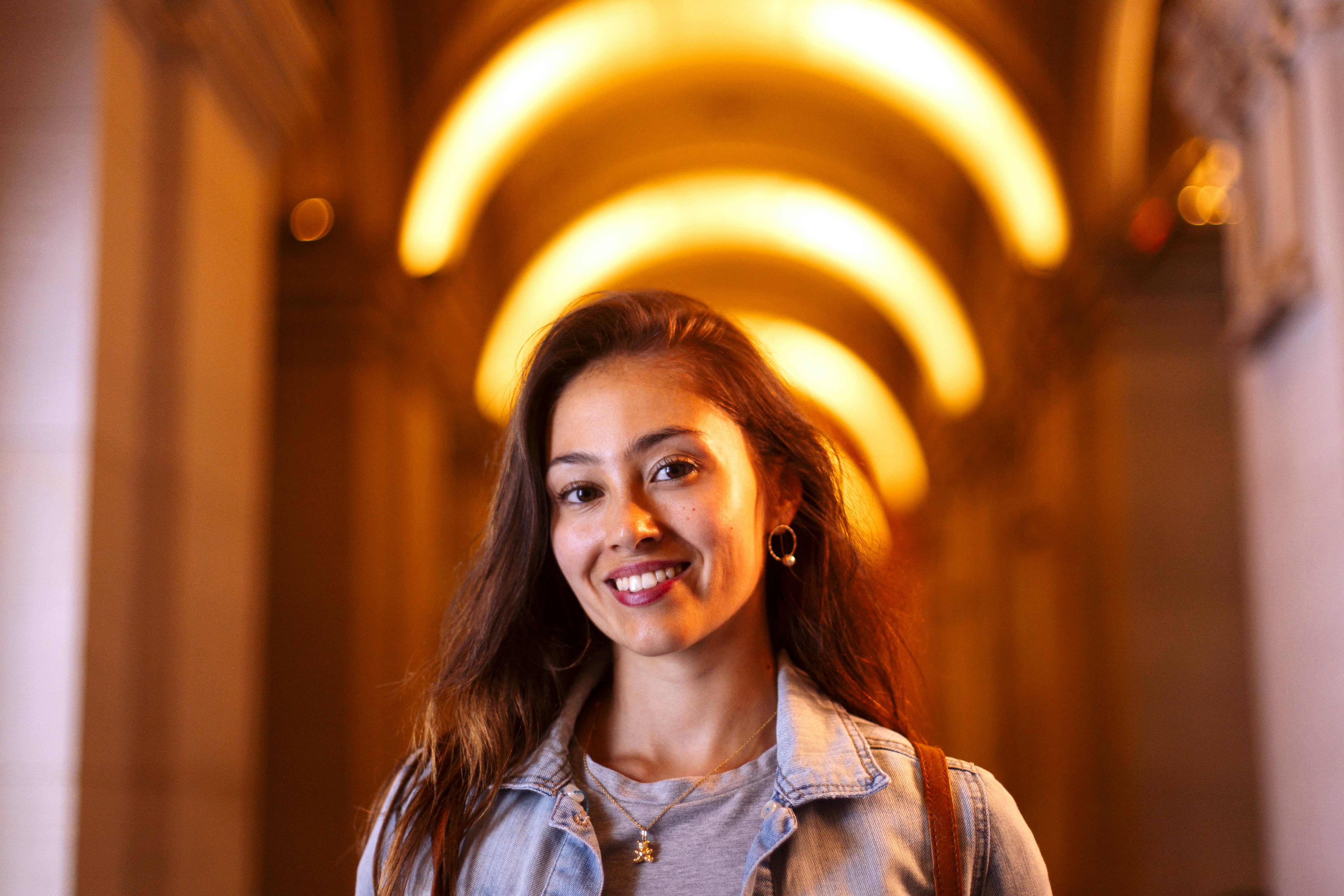 A cheerful young woman in a denim jacket poses in a warmly lit hallway with glowing arches, conveyin...