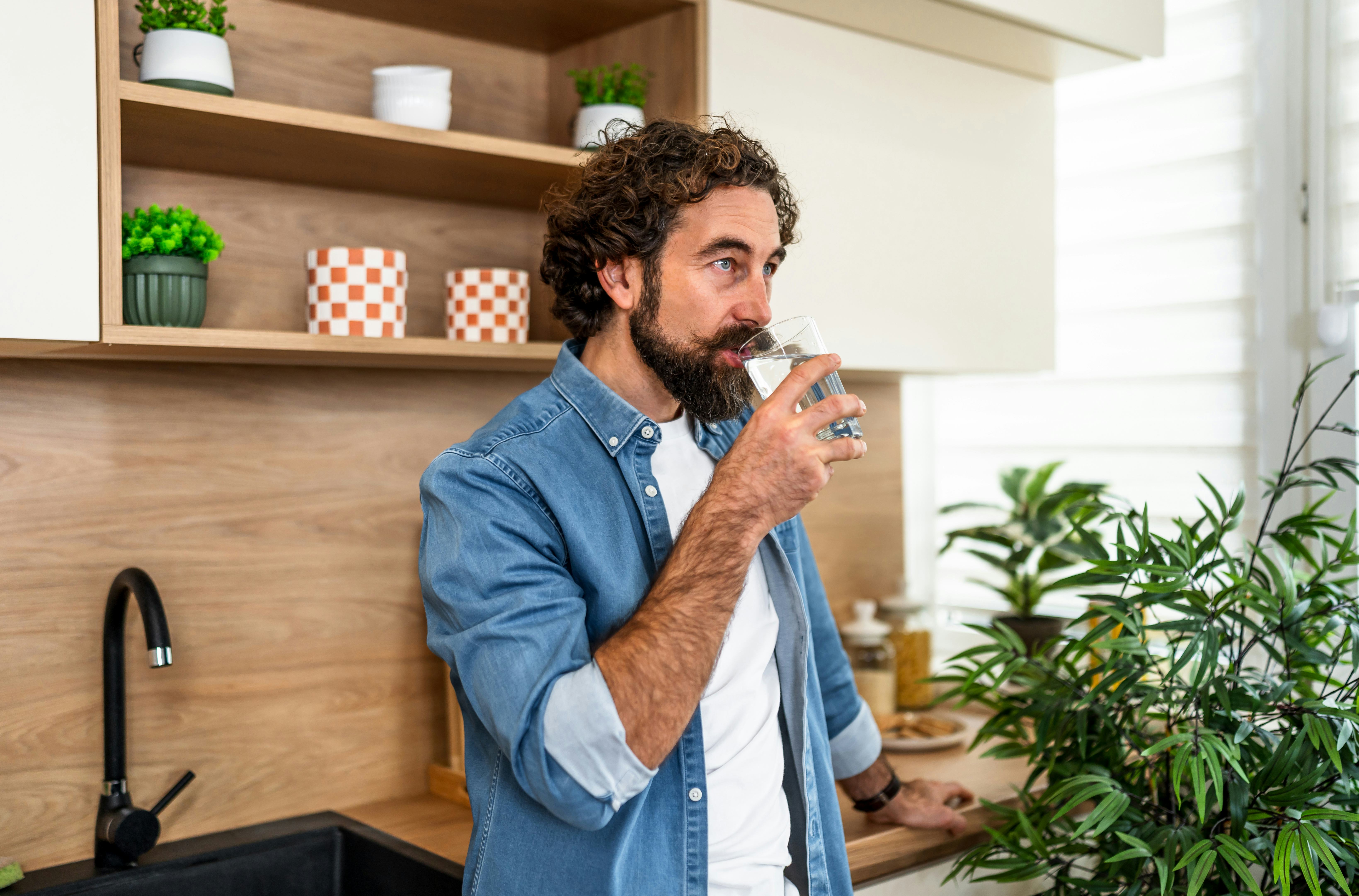 Man with a beard drinking water from a glass, standing in a contemporary kitchen at home, staying hy...