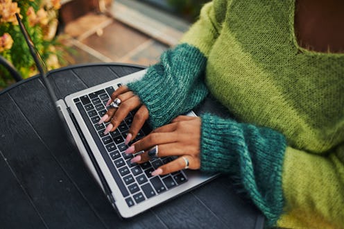 Close-up of female hands with rings typing on a laptop keyboard wearing a green and teal cozy sweate...