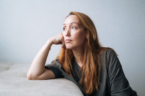 A melancholic adult woman gazes out of a window, lost in thought. Soft natural light highlights her ...