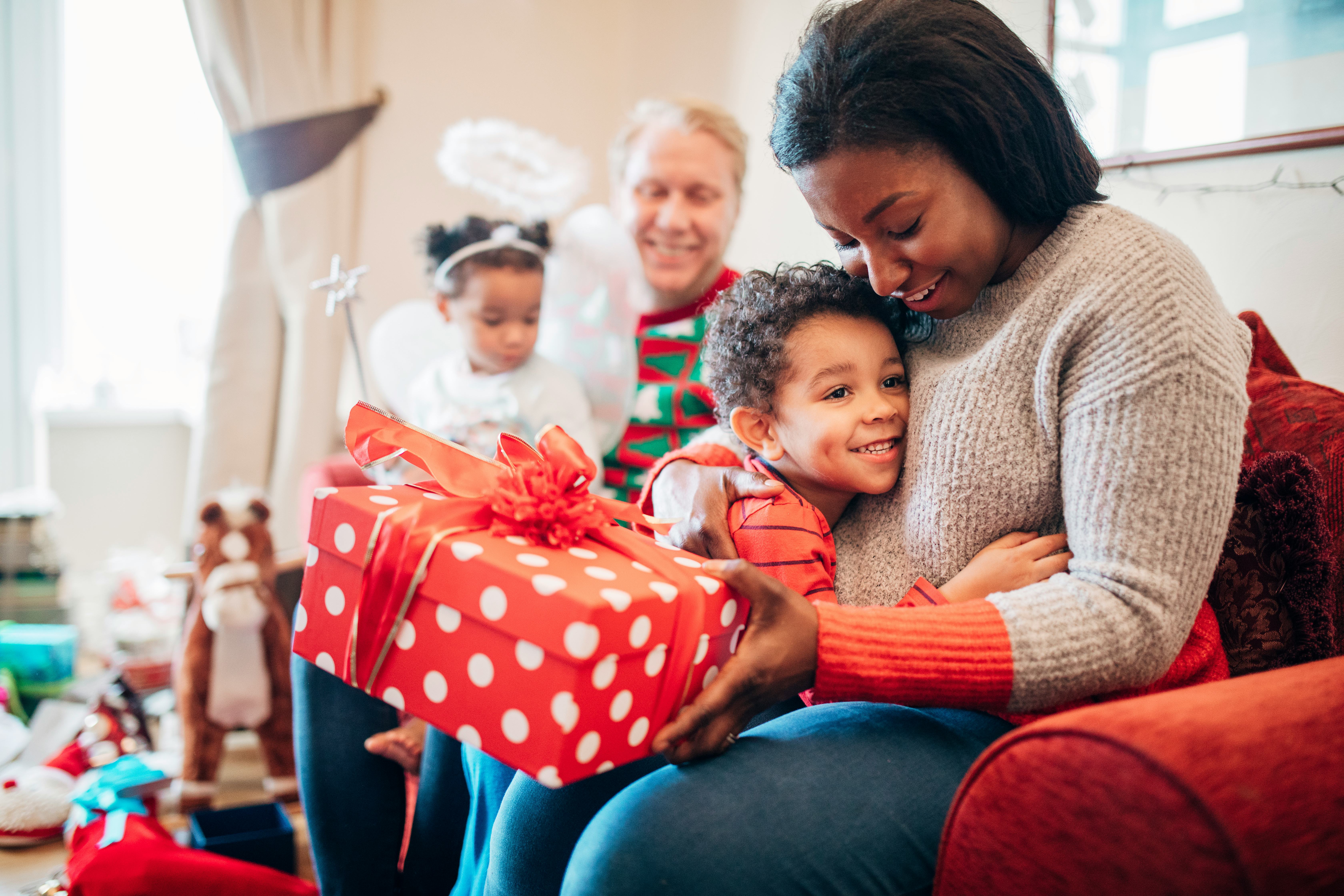 A little boy happily hugs his mother as she receives a gift from him on Christmas morning. His siste...