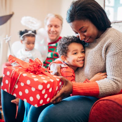 A little boy happily hugs his mother as she receives a gift from him on Christmas morning. His sister and father can be seen in the background.
