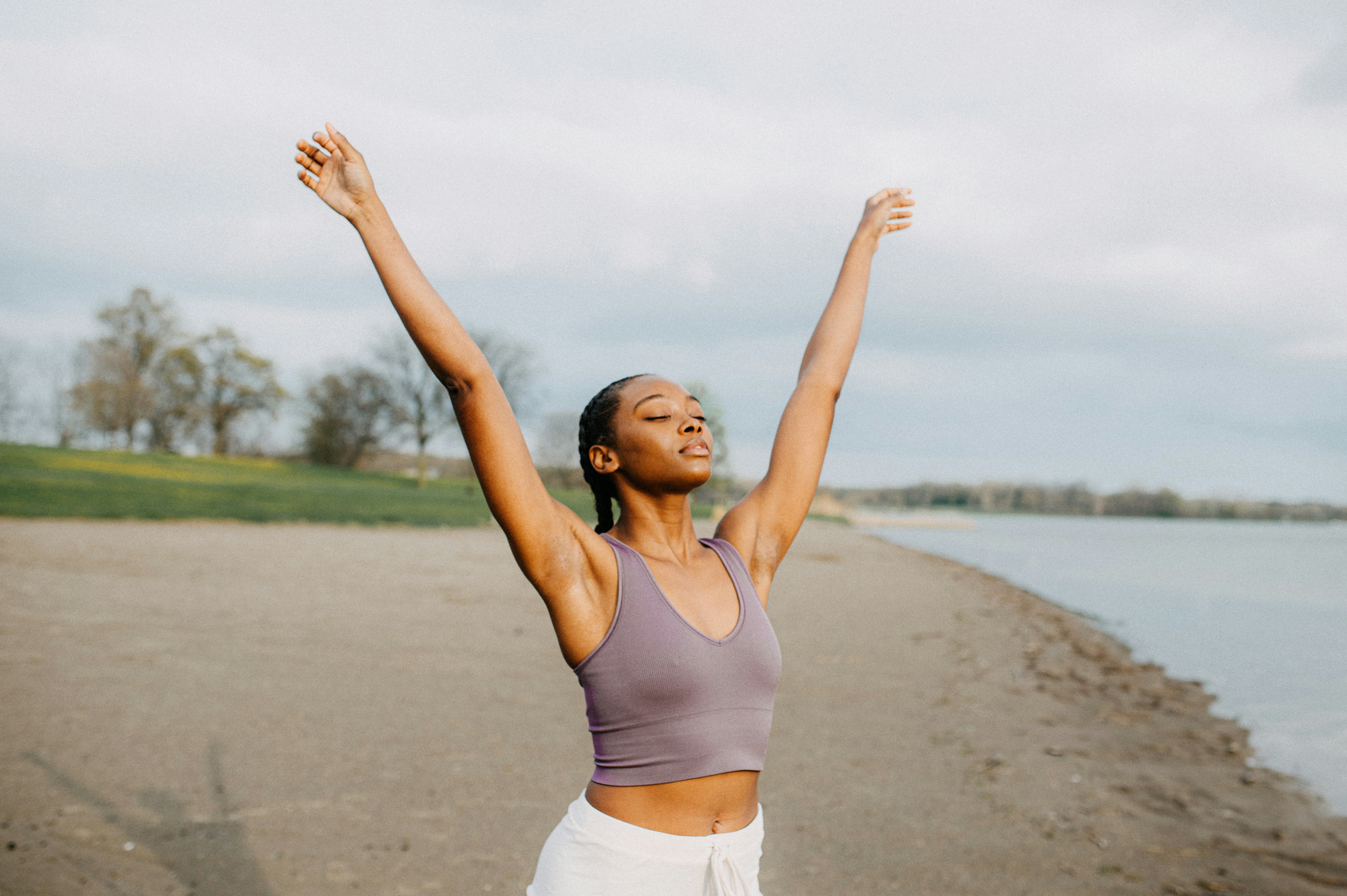 A woman stands on a beach with her arms raised, appearing serene and content. The sky is overcast, y...