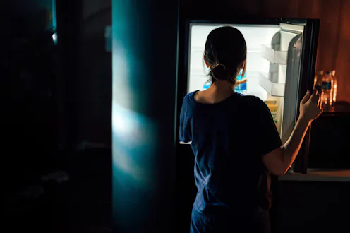 Rear view of young Asian woman standing in kitchen in the dark opening the fridge and look inside in...