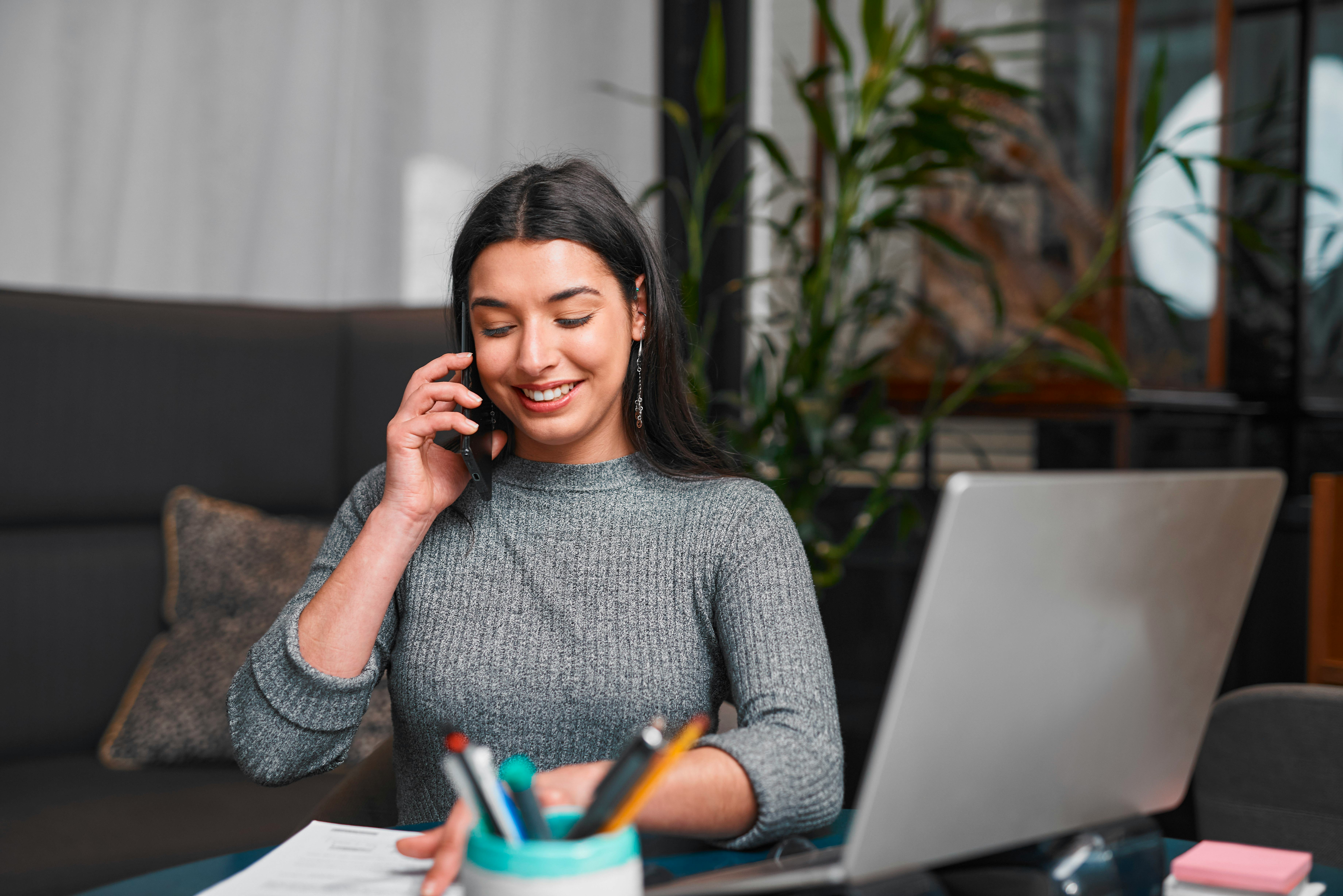 A focused professional woman chats on her mobile while working at a sleek desk with a laptop, papers...