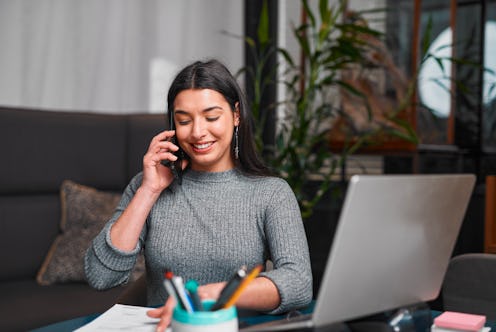 A focused professional woman chats on her mobile while working at a sleek desk with a laptop, papers...
