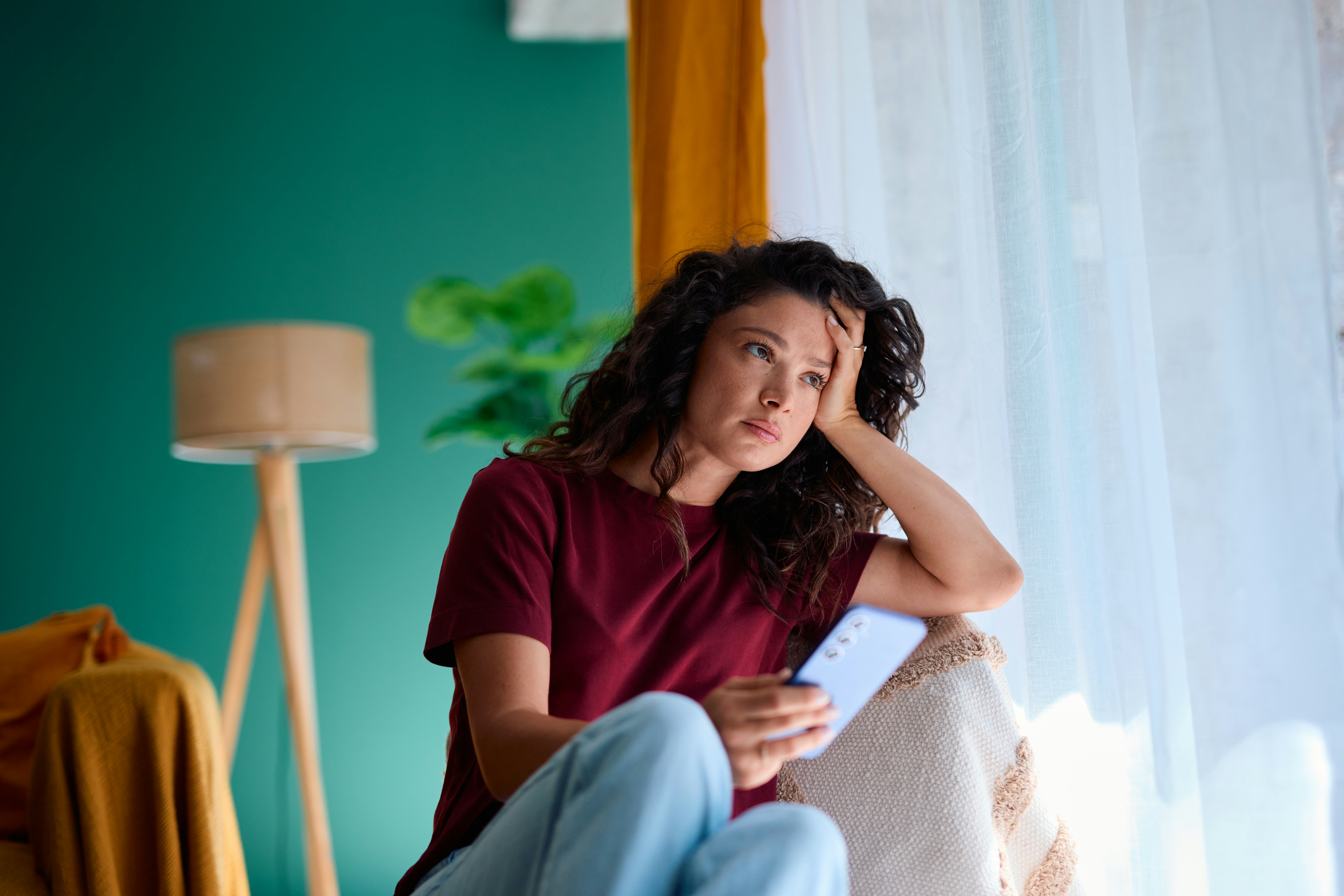 Worried young woman sitting by the window, holding a smartphone in her hand, lost in thought while c...