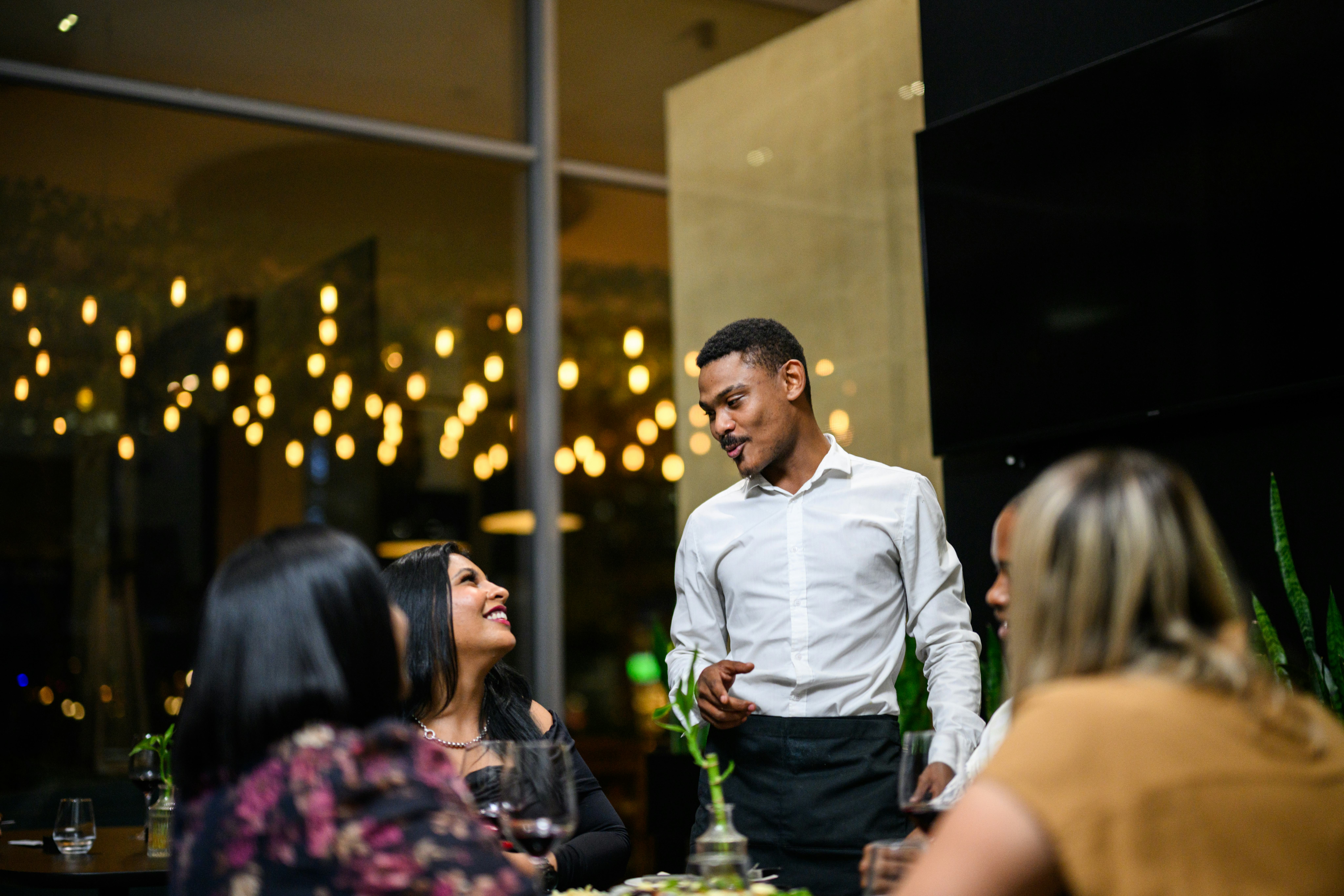Waiter talking to a group of friends enjoying an evening meal around a restaurant table in a luxury ...