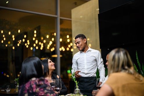 Waiter talking to a group of friends enjoying an evening meal around a restaurant table in a luxury ...
