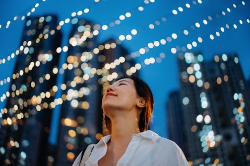 Low angle shot of beautiful young Asian woman standing against illuminated string lights in front of...