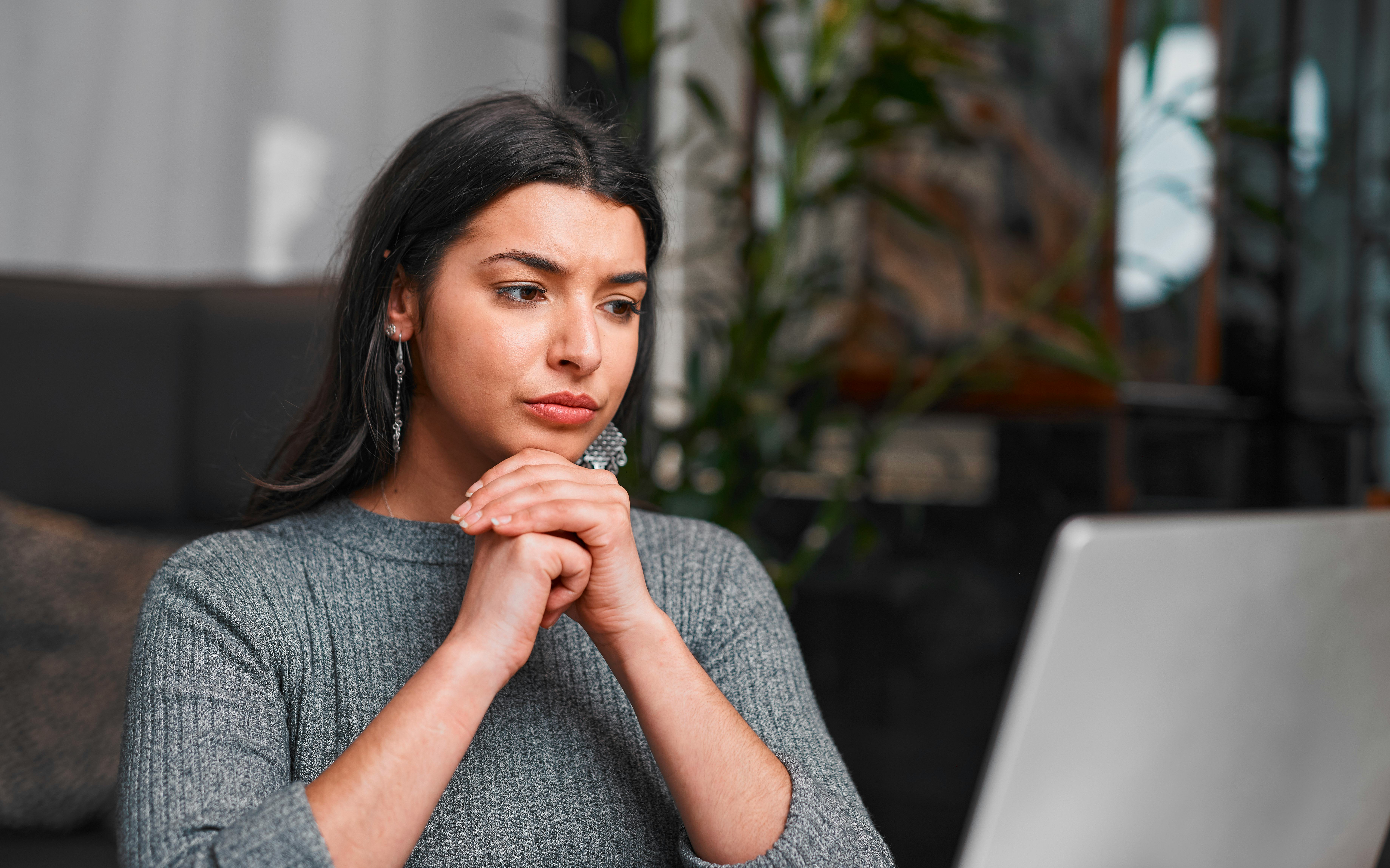 A thoughtful woman sits with clasped hands beside a laptop in a modern indoor space, with plants in ...