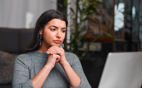 A thoughtful woman sits with clasped hands beside a laptop in a modern indoor space, with plants in ...