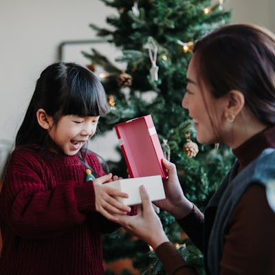 Loving Asian mother and cute little daughter enjoying opening Christmas presents together on Christmas morning, sharing the moment together. Decorated Christmas tree in background. Christmas lifestyle. Celebrating Christmas. Holiday and festive vibes
