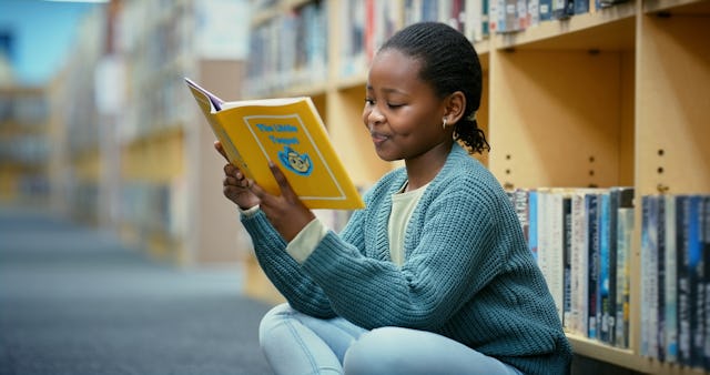 A smiling girl with braided hair wearing a sweater sits on the floor of a library reading a book.