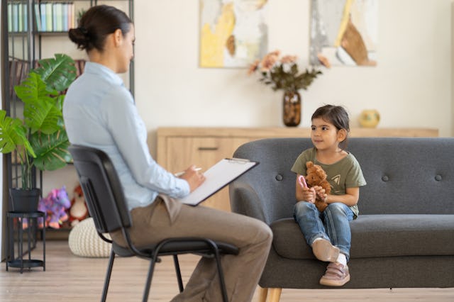 A child with dark hair in braids holds a teddy bear, sitting on a couch in a bright, well-appointed ...
