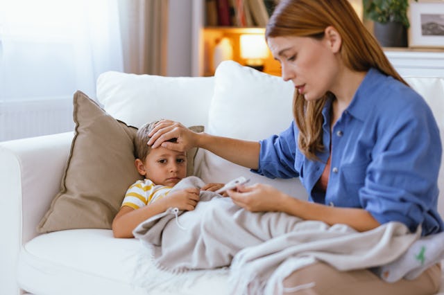 A mother attentively cares for her sick child laying on a white couch, covering him with a blanket a...
