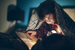 Little girl reading a book under a blanket in the dark