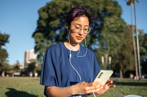 A young person listens to music with earphones outdoors in a park setting, engaging with their phone...