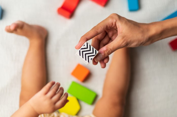 mother helping 6-9 months old baby playing with multi colored wooden block toys, sitting on a white ...