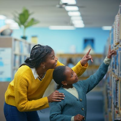 A librarian in a yellow sweater helps a child choose a book on a high shelf.