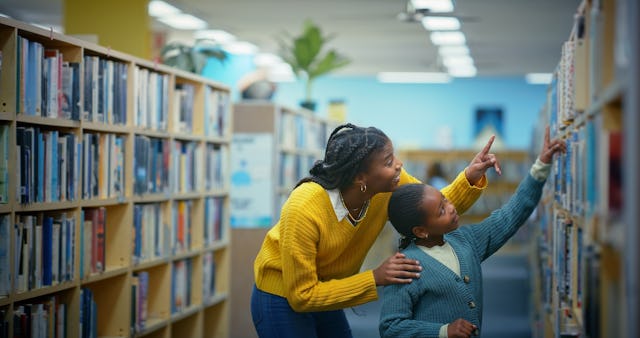 A librarian in a yellow sweater helps a child choose a book on a high shelf.
