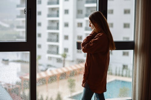 Young woman looking over window pane holding coffee. Thoughtful woman thinking and looking away whil...