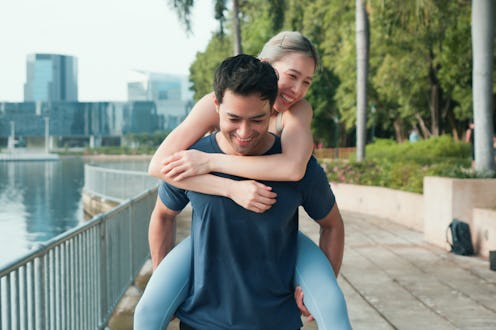 A smiling man gives his partner a piggyback ride along a scenic urban lakeside path. Lush greenery, ...