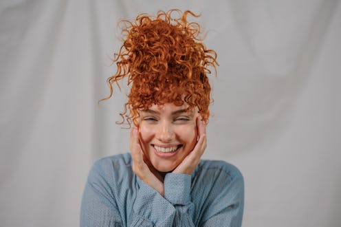 Portrait of a cheerful young woman with curly red hair, smiling and touching her face with her hands