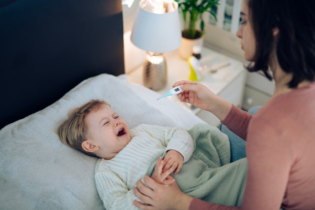 A mother gently checks her crying toddler's temperature at home, offering comfort as the child sits ...
