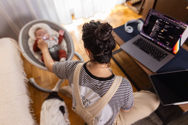A mother using her laptop while her baby sleeps soundly in a nearby swing.